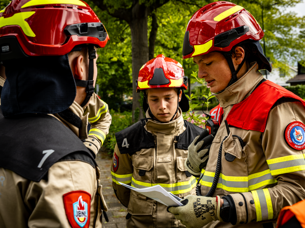 Een ploeg jeugdbrandweerleden in uitrukpak met helm op staat buiten rond de bevelvoerder, die de opdracht voorleest tijdens een jeugdbrandweerwedstrijd