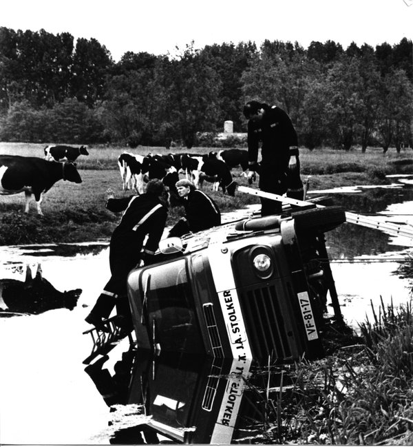 Historische zwart wit foto van brandweervoertuig in de sloot. Brandweermensen zitten op het voertuig.