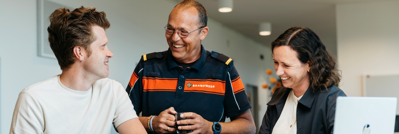 Drie mensen staan lachend aan een bureau in een kantooromgeving