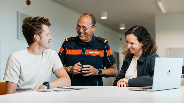 Drie mensen staan lachend aan een bureau in een kantooromgeving