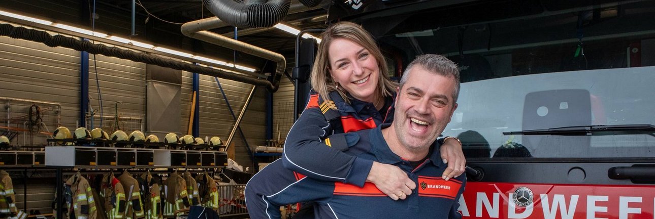 Brandweervrouw op rug van brandweerman lachend in de camera, voor een brandweervoertuig op brandweerpost