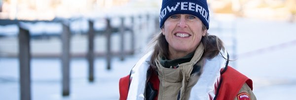 Vrouw met muts op in brandweerbluspak staat lachend in de sneeuw