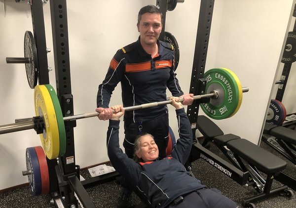 2 brandweermensen poseren lachend voor de camera terwijl 1 collega aan het bankdrukken is