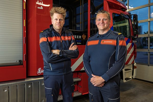 Twee brandweermensen, Gerard en Chris, staan voor een brandweervoertuig van de post Zuilen.