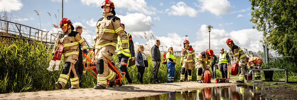 Leden van jeugdbrandweer lopen in uitrukpak met slangenkar en brandweerslangen langs regenplas tijdens een oefening