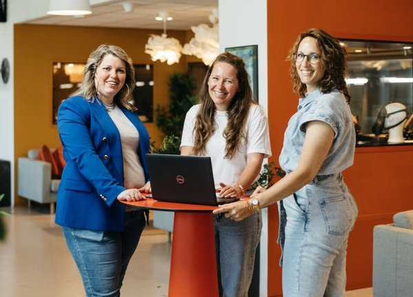 Drie vrouwen staan lachend aan een statafel in een kantooromgeving