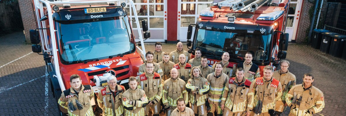 twee brandweervoertuigen achter een brandweerpost met collega's