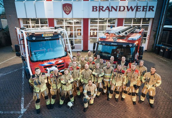 twee brandweervoertuigen achter een brandweerpost met collega's